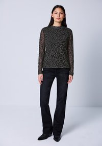 Kvinde står iført en sort langærmet blomster mesh-top, sorte lige benbukser og sorte spidse støvler mod en grå baggrund.