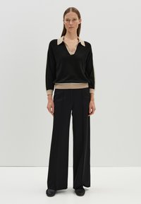 Maglione nero a maglia con colletto e polsini beige, caratterizzato da un design a V. Abbinato a pantaloni neri a gamba larga e scarpe scure.