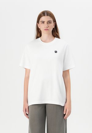 Il modello indossa una semplice maglietta bianca decorata con un piccolo logo a forma di fiore nero, abbinata a pantaloni grigi a gamba larga, il tutto su uno sfondo bianco.