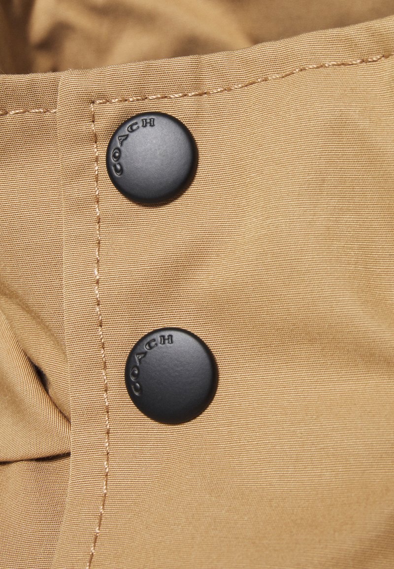 Deux boutons noirs mats avec le texte "COACH" sur un tissu beige avec des coutures visibles, faisant partie du détail de fermeture d'un vêtement.