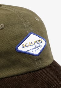 Gorra verde oliva con una visera de pana marrón. Presenta un parche en forma de diamante en blanco y azul etiquetado "SCALPERS simplicité." Tejido texturizado.