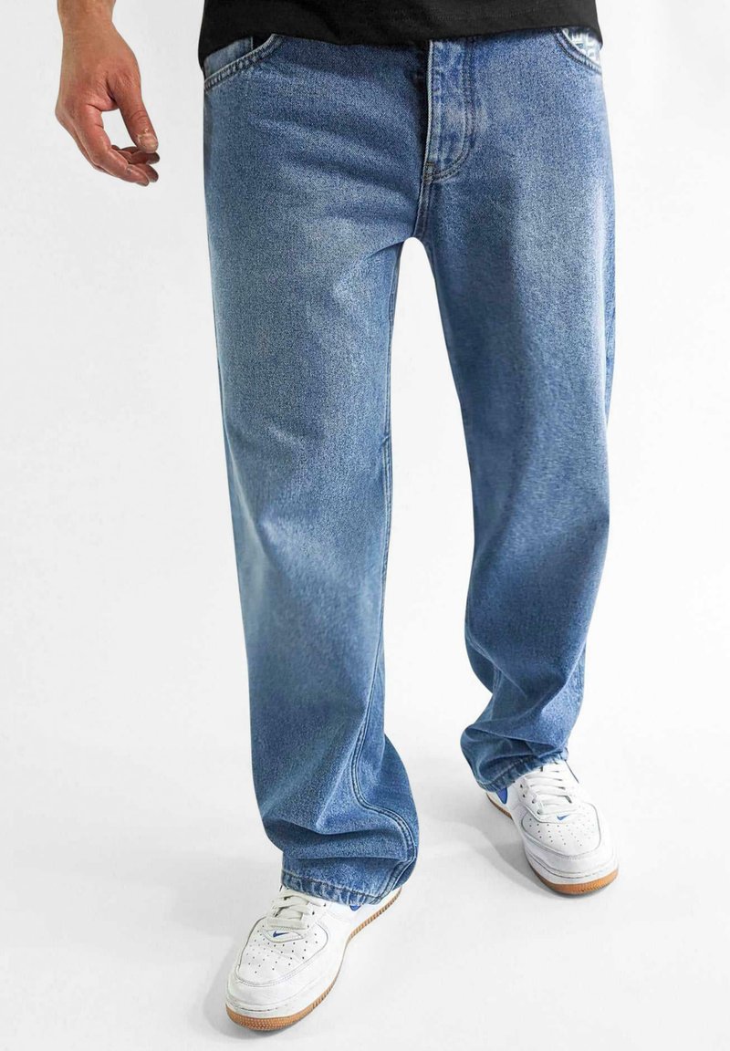 Jeans in denim blu chiaro con una vestibilità comoda, caratterizzati da cinque tasche e un leggero sbiadimento, abbinati a sneakers bianche su uno sfondo neutro.