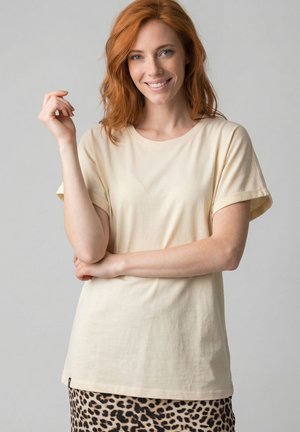 Lächelnde rothaarige Frau in cremefarbenem Kurzarmshirt und Leopardenmuster-Rock steht mit verschränkten Armen und erhobener Hand vor grauem Hintergrund.