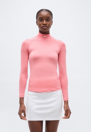 Kobieta ubrana w różowy, długi rękaw z golfem i białą spódnicę, stojąca na jednolitym jasnym tle.