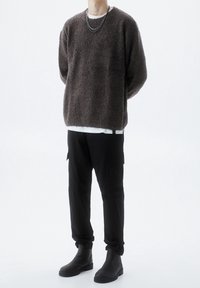 Maglione marrone con texture, dal taglio morbido, indossato sopra una camicia bianca. I pantaloni cargo neri e gli stivaletti completano l'outfit.