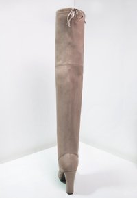 Botte pour femme en suède beige, haute, avec un talon élevé et une fermeture à lacets en haut, vue de dos sur une surface blanche.