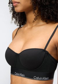 Γυναίκα με σγουρά μαλλιά που φοράει μαύρο σουτιέν Calvin Klein με λεπτές τιράντες και ελαστική ταινία με το λογότυπο, εμφανιζόμενη από το λαιμό έως το μέσο της κοιλιάς.