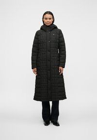 Schwarzer, gesteppter langer Mantel mit Kapuze, ausgestattet mit einem Frontdruckknopfverschluss und rechteckigen Mustern. Trägt dazu dunkle Jeans und glänzende schwarze Schuhe.