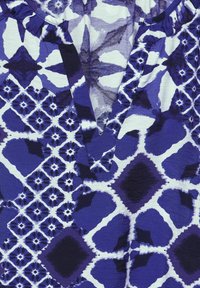 Lila und weiße gemusterte Stoff mit V-Ausschnitt und Rüschendetail. Das Design umfasst florale und geometrische Formen mit einer strukturierten Oberfläche.