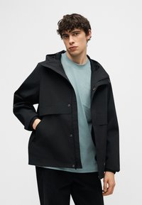 Νέος άνδρας με καστανά σγουρά μαλλιά, φορώντας μαύρο μπουφάν με κουκούλα πάνω από ανοιχτό μπλε μπλουζάκι, ποζάρει με το ένα χέρι στην τσέπη του μπουφάν.