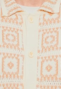 Πουλόβερ σε μπεζ και ροδακινί χρώμα με γεωμετρικά τετράγωνα σχέδια και δύο εμφανή κουμπιά πάνω σε λευκό τοπ.