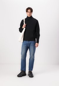 Veste zippée noire avec un col montant, associée à un jean en denim bleu clair et des baskets noires. Modèle tenant un sac fourre-tout blanc.