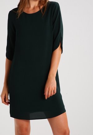 Frau trägt ein dunkelgrünes, knielanges Kleid mit halblangen Ärmeln und vorderen Schlitzen, steht vor einem einfarbigen hellen Hintergrund.