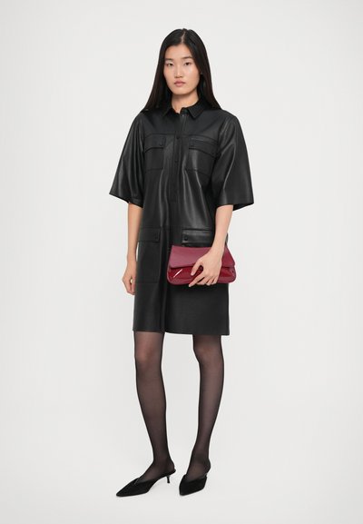 Γυναίκα με κοντό μανίκι μαύρο δερμάτινο φόρεμα, μαύρες διαφανείς καλσόν, μαύρες κομψές γόβες, κρατώντας ένα γυαλιστερό κόκκινο τσαντάκι σε απλό λευκό φόντο.