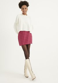 Top bianco a maniche lunghe e volant abbinato a una mini gonna a quadri rossi. La modella indossa stivali beige alti fino al ginocchio con cerniere laterali nere. Collant neri.
