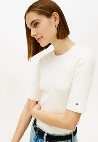 Cienki, krótki top w kolorze kremowym z okrągłym dekoltem, z małym logotypem marki na rękawie, zestawiony z jasnoniebieskimi dżinsami.