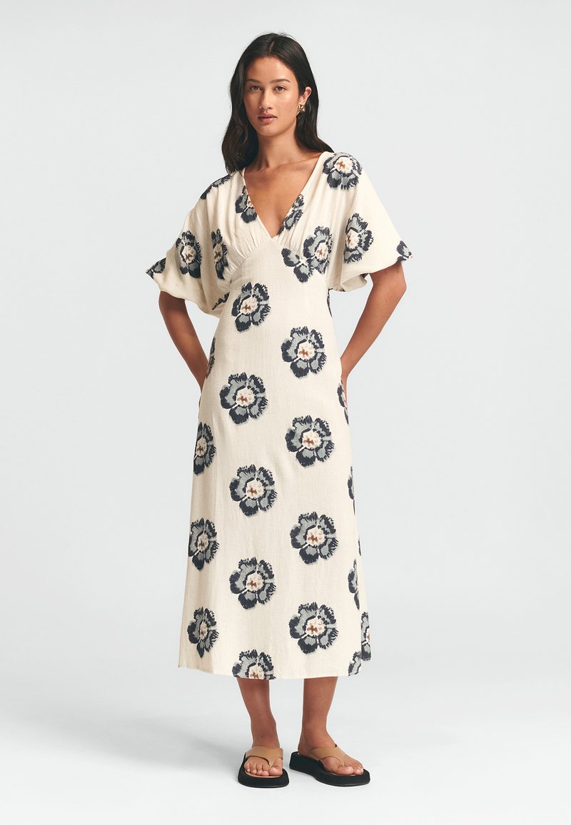 Krämfärgad blommig klänning med marinblå blommönster, V-ringning och korta vida ärmar. Bärs med svarta platta sandaler. Texturen verkar lätt och luftig.