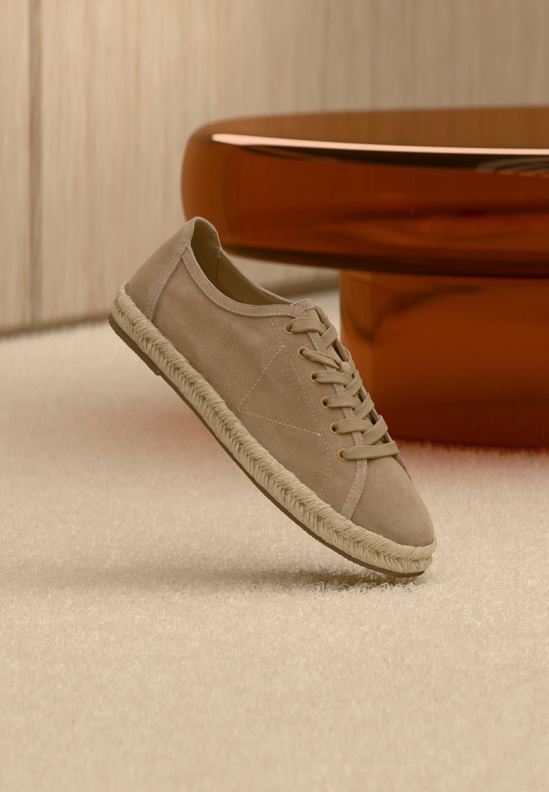 Baskets beiges en daim à lacets avec une semelle tressée flottant au-dessus d'un tapis crème, avec une table moderne courbée de couleur ambre en arrière-plan.
