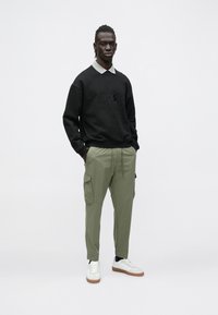 Μαύρο φούτερ με λογότυπο "BOSS", γκρι πουκάμισο με γιακά από κάτω, παντελόνι cargo σε ελαιί απόχρωση και λευκά αθλητικά παπούτσια με καφέ σόλες.