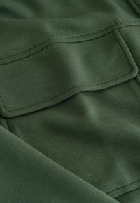 Close-up of dark green fabric with a rectangular stitched pocket flap and visible seam detailing.