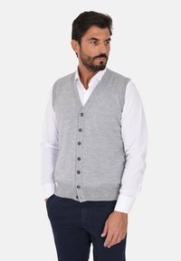 Gilet di lana grigia con scollatura a V, dotato di chiusura con bottoni e orlo a coste discreto. Indossato sopra una camicia bianca, abbinato a pantaloni scuri.