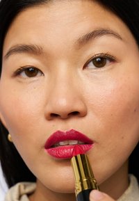 Lipstick in een gouden en zwarte tube, heldere roze aangebracht op de lippen, gladde textuur, glanzende afwerking, close-up van het gezicht. Bruine ogen en een lichte huid.