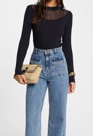 Woman wearing a black ribbed long-sleeve top with sheer striped neckline, high-waisted blue jeans, gold bracelet, and holding a woven clutch bag.