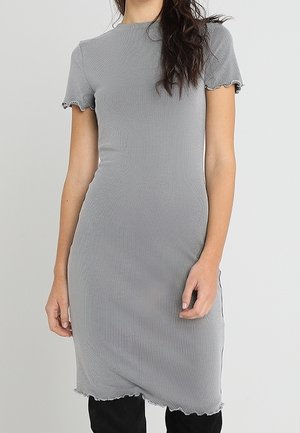 Kvinde iført en tætsiddende, ribbet grå kjole med korte flæseærmer og -kant, stående mod en ensfarvet lys baggrund.