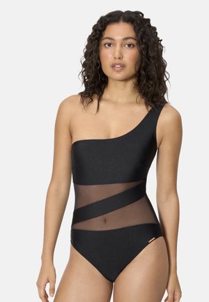 Mujer con un traje de baño negro de un solo hombro con paneles diagonales transparentes en la zona media, de pie contra un fondo claro y sencillo.