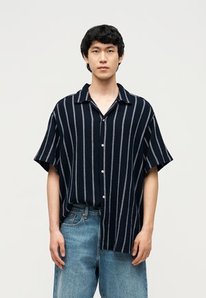 Jeune homme debout, face à l'avant, portant une chemise à manches courtes bleu marine à rayures verticales blanches et un jean bleu clair, sur un fond uni.