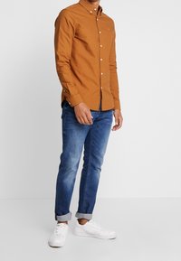 Bruine lange mouwen knoopshirts, katoen stof, slim fit; gecombineerd met opgerolde mouw blauwe jeans en witte sneakers. Knoopdetails op het shirt.