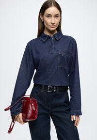 Mørk denimskjorte med lange ærmer, krave og brystlomme, kombineret med højtaljede mørke jeans. Holder en lille, rød læderhåndtaske.