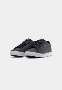 Une paire de baskets noires à lacets avec des semelles blanches et une discrète bordure rouge, marquées "Tommy Hilfiger" sur le côté et la languette, sur un fond blanc.