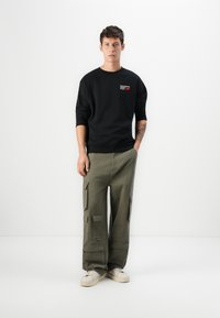 Black sweatshirt with text on the chest, paired with olive green cargo pants featuring multiple pockets. Light-colored sneakers complete the look.