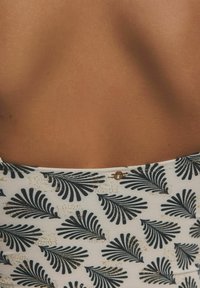 Witte stof met een marineblauw palmbladpatroon en gouden accenten, wat een textuur aan het geheel geeft. Glad huid is zichtbaar rond het kledingstuk.
