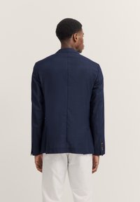 Blazer blu navy con tessuto testurizzato, colletto a rovescio e polsini a quattro bottoni, esposto da dietro su uno sfondo chiaro.