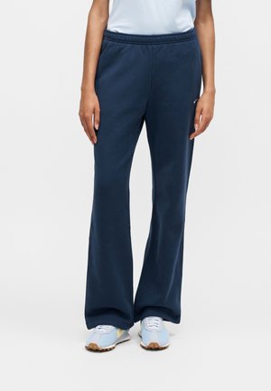 Personne portant un pantalon de survêtement bleu marine à jambes larges et des baskets bleu clair et blanches, debout sur un fond blanc uni.
