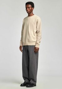 Bēša adīta džempera ar apaļu kakla izgriezumu, kopā ar plašām pelēkām svītrainām biksēm un melniem apaviem. Džemperim ir teksturēts audums.