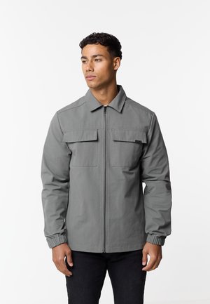 Mężczyzna ubrany w szarą kurtkę na zamek błyskawiczny z klapami kieszeni na piersi i elastycznymi mankietami, stojący na tle białego, jednolitego tła, patrzący w lewo.