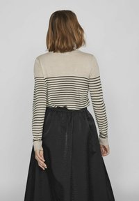 Osoba o włosach do ramion nosi pasiasty sweter w beżowe i czarne paski połączony z czarną spódnicą, widziana od tyłu.