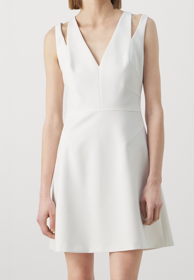 Femme portant une robe blanche sans manches avec un col en V profond et une taille cintrée, debout devant un fond clair uni.
