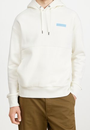 Hoodie blanc en tissu doux, avec une poche kangourou et un patch logo bleu sur la poitrine. Poignets et taille côtelés.