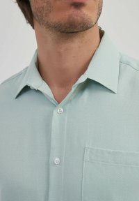 Chemise boutonnée vert clair avec un tissu texturé, un col pointu et une poche poitrine. Boutons blancs sur le devant.