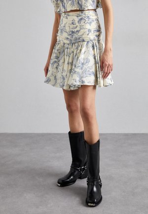Jupe à imprimé floral en crème et bleu, avec un ourlet à volants et des détails froncés. Associée à des bottes noires montantes avec des accents métalliques.