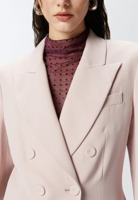 Svijetlopinkasta dvostruko kopčana blejzerica od glatkog materijala, s reverima i dva velika gumba. Nosi se preko prozirne, točkaste boje tamnocrvene bluze.