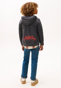 Mørk grå hættetrøje med rød tekst på ryggen, med en lomme foran. Kombineret med blå jeans og beige sneakers. Ensfarvet baggrund.