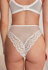 Balts mežģīņu apakšveļas komplekts ar caurspīdīgu audumu, viļņotām mežģīņu malām un augstu griezumu. Aizmugurē regulējams āķa aizdares elementu.