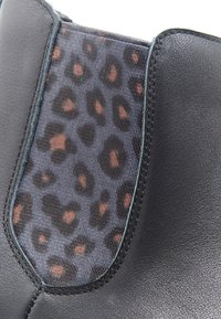 Botín negro de piel con panel lateral elástico texturizado de patrón de leopardo que presenta manchas marrones y negras, con costuras a lo largo de los bordes.