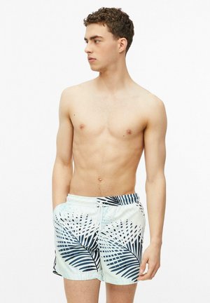 Jeune homme aux cheveux bouclés portant un short de bain blanc avec des motifs de feuilles de palmier bleu foncé, debout les mains dans les poches, regardant vers la gauche.