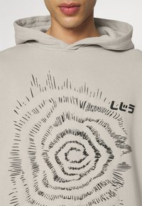 Sweat à capuche beige clair avec un graphisme en spirale noir, au design texturé. Petit texte sur le côté, mettant en avant un style minimaliste.